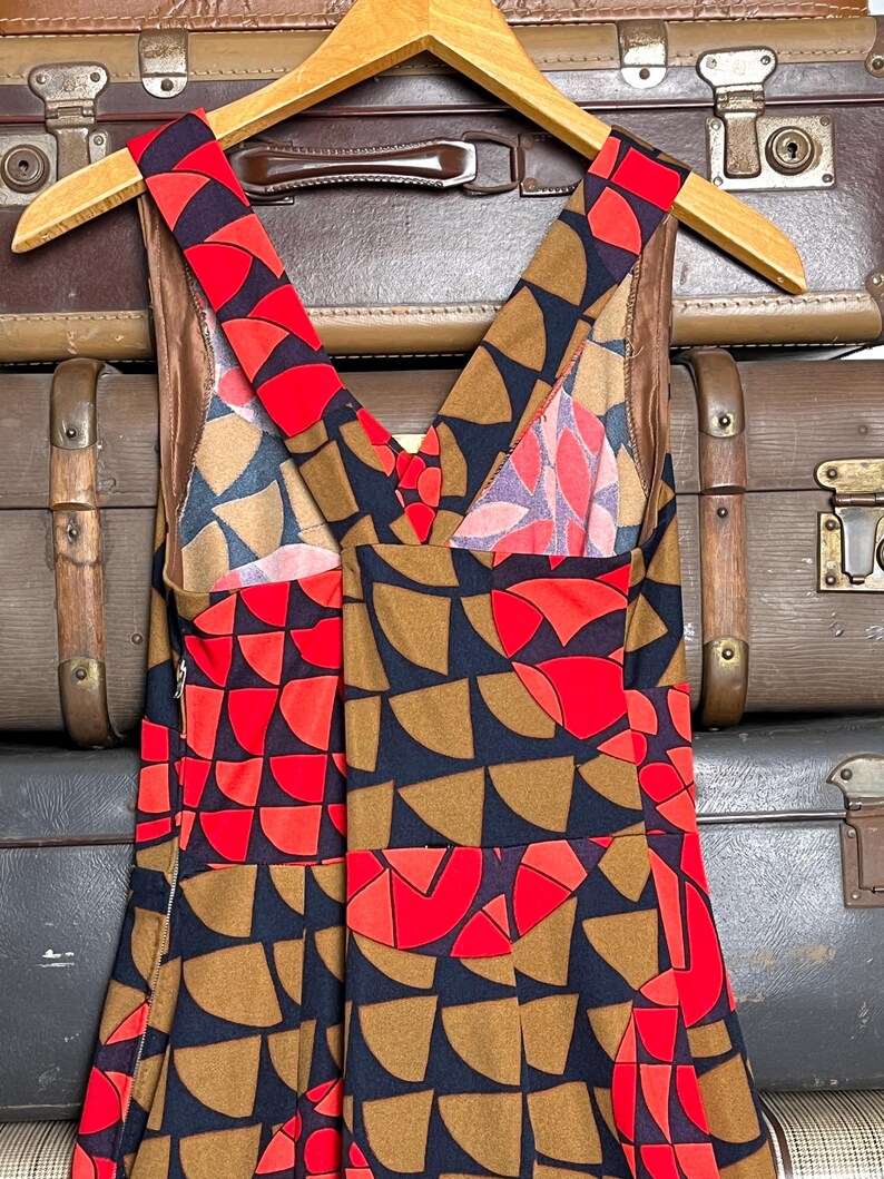 May include: A sleeveless dress featuring a geometric pattern of semicircles in red, tan, and black. The garment has a V-neckline, wide shoulder straps, and a side zipper closure. The interior is lined with a solid brown fabric, and the dress is displayed on a wooden hanger.