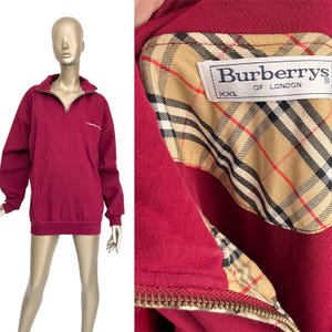 May include: A burgundy sweatshirt with a quarter-zip neckline. The collar lining features a classic plaid pattern. The Burberrys of London label is visible. The sweatshirt is displayed on a mannequin.