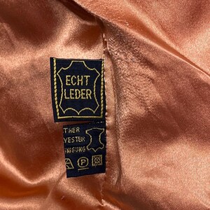 May include: Close-up of a peach-colored satin fabric with a black and gold tag. The tag reads "ECHT LEDER" in gold lettering, indicating genuine leather. Additional text and care symbols are visible on a smaller tag.