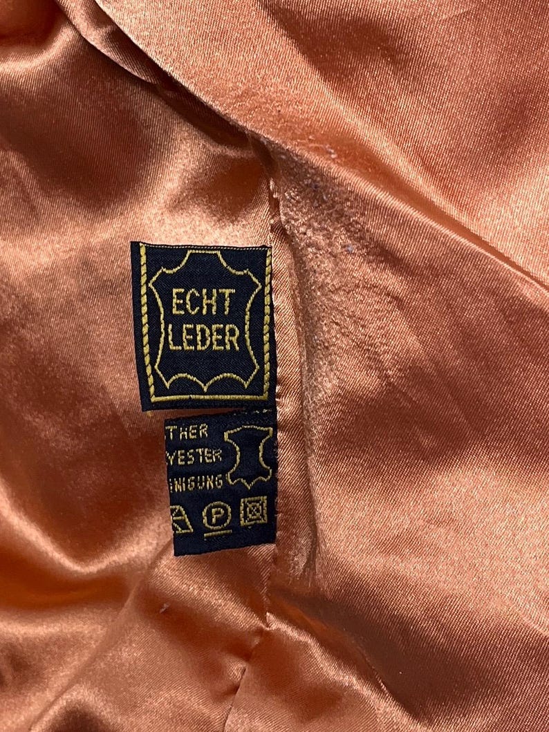 May include: Close-up of a peach-colored satin fabric with a black and gold tag. The tag reads "ECHT LEDER" in gold lettering, indicating genuine leather. Additional text and care symbols are visible on a smaller tag.