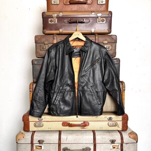 May include: Black leather jacket with a zip-up front, two front pockets, and a collared neckline. The jacket is hanging on a wooden hanger and is displayed against a backdrop of vintage suitcases. The jacket's interior lining is orange.
