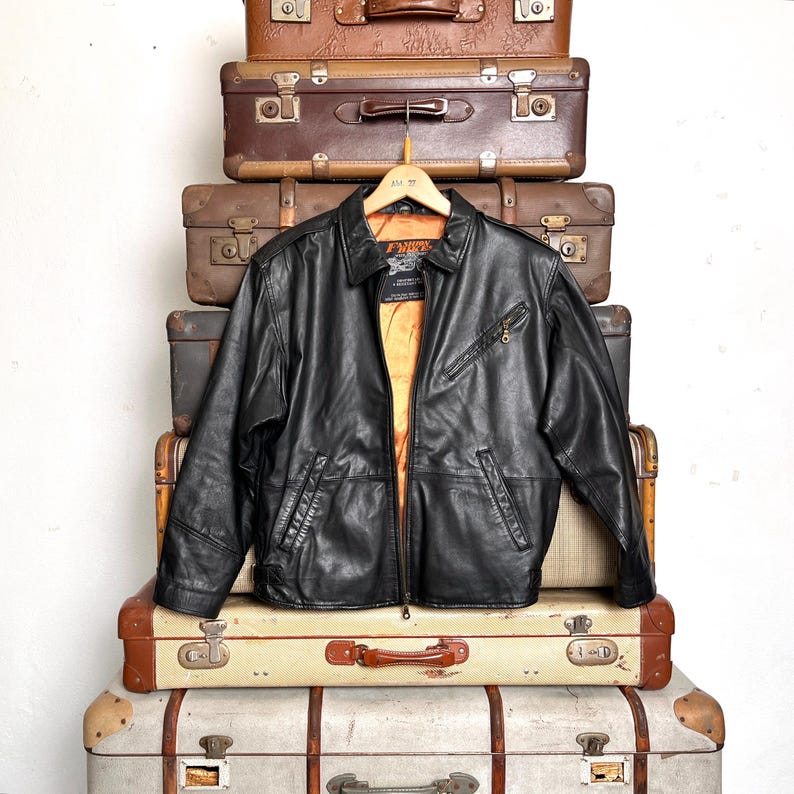 May include: Black leather jacket with a zip-up front, two front pockets, and a collared neckline. The jacket is hanging on a wooden hanger and is displayed against a backdrop of vintage suitcases. The jacket's interior lining is orange.