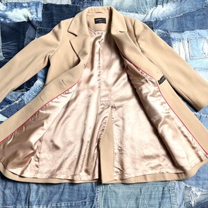 May include: A beige overcoat with a satin lining and red trim. The coat is open, revealing the interior lining. The coat has a classic lapel and button closure. The coat is laid out on a denim patchwork background.