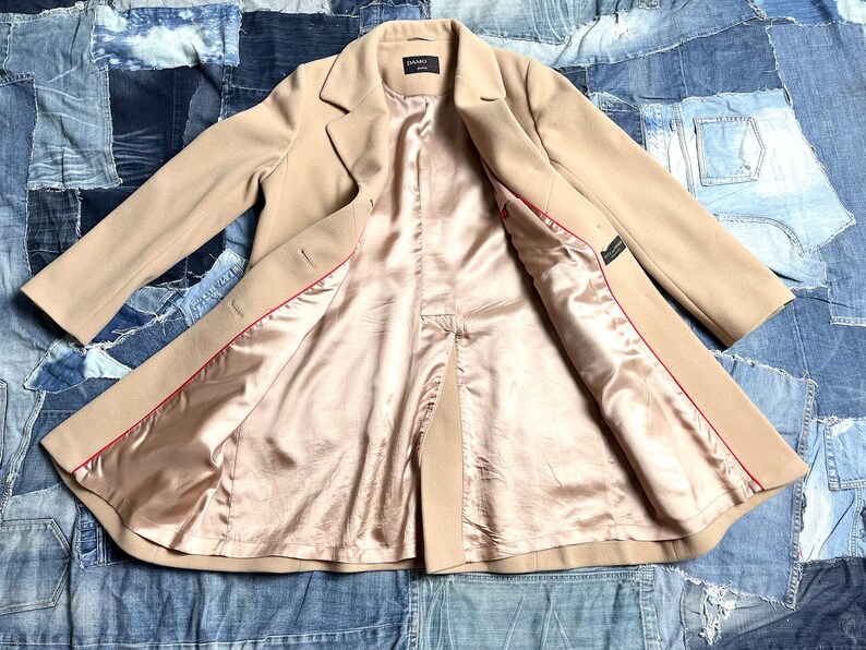 May include: A beige overcoat with a satin lining and red trim. The coat is open, revealing the interior lining. The coat has a classic lapel and button closure. The coat is laid out on a denim patchwork background.