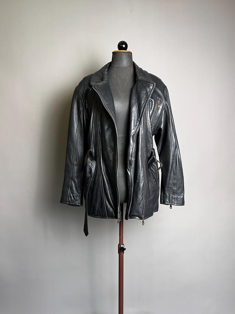 May include: A black leather jacket with a notched collar, zipper closure, and belt detail. The jacket is displayed on a mannequin and has a classic, vintage style. The jacket has a shiny finish and appears to be in good condition.