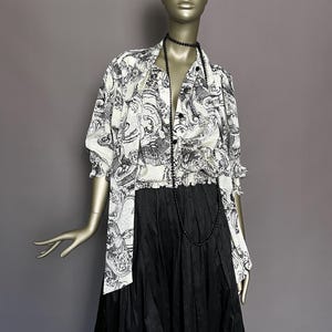 Puede incluir: Un maniquí exhibe una falda negra y una blusa estampada. La blusa presenta un diseño en blanco y negro, una botonadura frontal y mangas con volantes. Un collar de cuentas negras cuelga alrededor del cuello del maniquí.