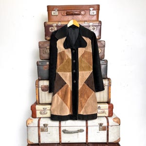 Peut inclure: Un manteau patchwork vintage dans les tons marron, beige et noir est suspendu à un cintre en bois. Le manteau présente un motif géométrique et est boutonné. Le manteau est présenté sur un fond de valises vintage empilées.