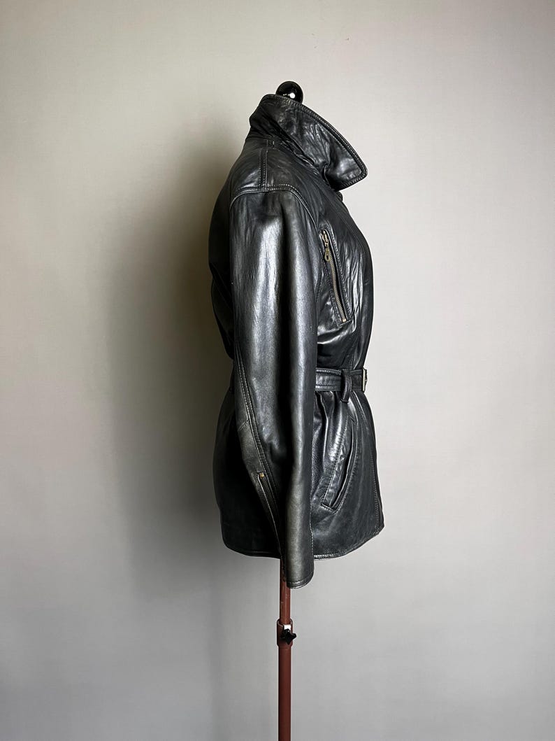 May include: Black leather jacket with a belted waist and a high collar. The jacket has a zipper closure and two front pockets. The jacket is displayed on a mannequin.
