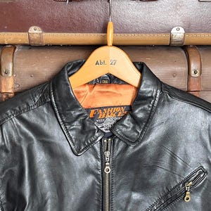 May include: Black leather jacket with a collared neckline, a zipper closure, and a zippered chest pocket. The jacket is hanging on a wooden hanger with the text "Abt. 27". The jacket has the text "Fashion Bikes Week Sport" on the label.