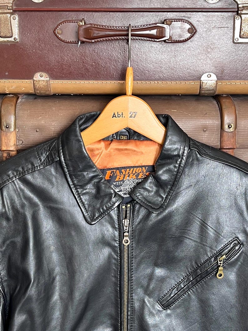 May include: Black leather jacket with a collared neckline, a zipper closure, and a zippered chest pocket. The jacket is hanging on a wooden hanger with the text "Abt. 27". The jacket has the text "Fashion Bikes Week Sport" on the label.