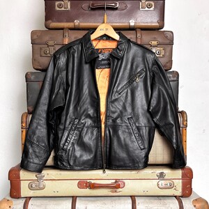 May include: A black leather jacket with a zip closure and a collared neckline hangs on a wooden hanger. The jacket has a bright orange lining and is displayed against a backdrop of vintage suitcases.