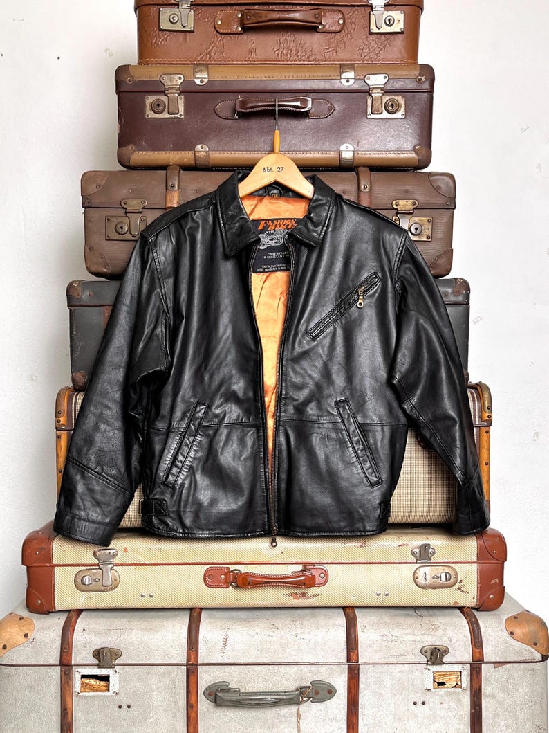 May include: A black leather jacket with a zip closure and a collared neckline hangs on a wooden hanger. The jacket has a bright orange lining and is displayed against a backdrop of vintage suitcases.