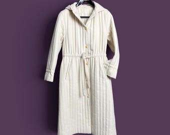 May include: A long-sleeved, off-white quilted coat featuring vertical stitching, a hood, and a front button closure. The garment includes a drawstring waist with a tie belt, two side pockets, and a structured collar, presented on a black hanger.
