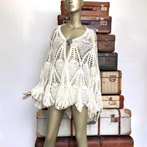May include: Off-white, open-weave crochet top with bell sleeves and a V-neckline. The top features a decorative clasp at the bust and a scalloped hem. The garment is displayed on a mannequin.