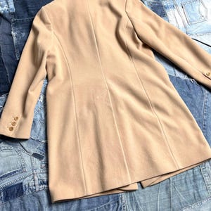 May include: A tan-coloured blazer with a classic design. The blazer features a notched lapel, long sleeves with buttoned cuffs, and a tailored fit. The back of the blazer has vertical seams for a structured look. The garment is made of a smooth fabric.