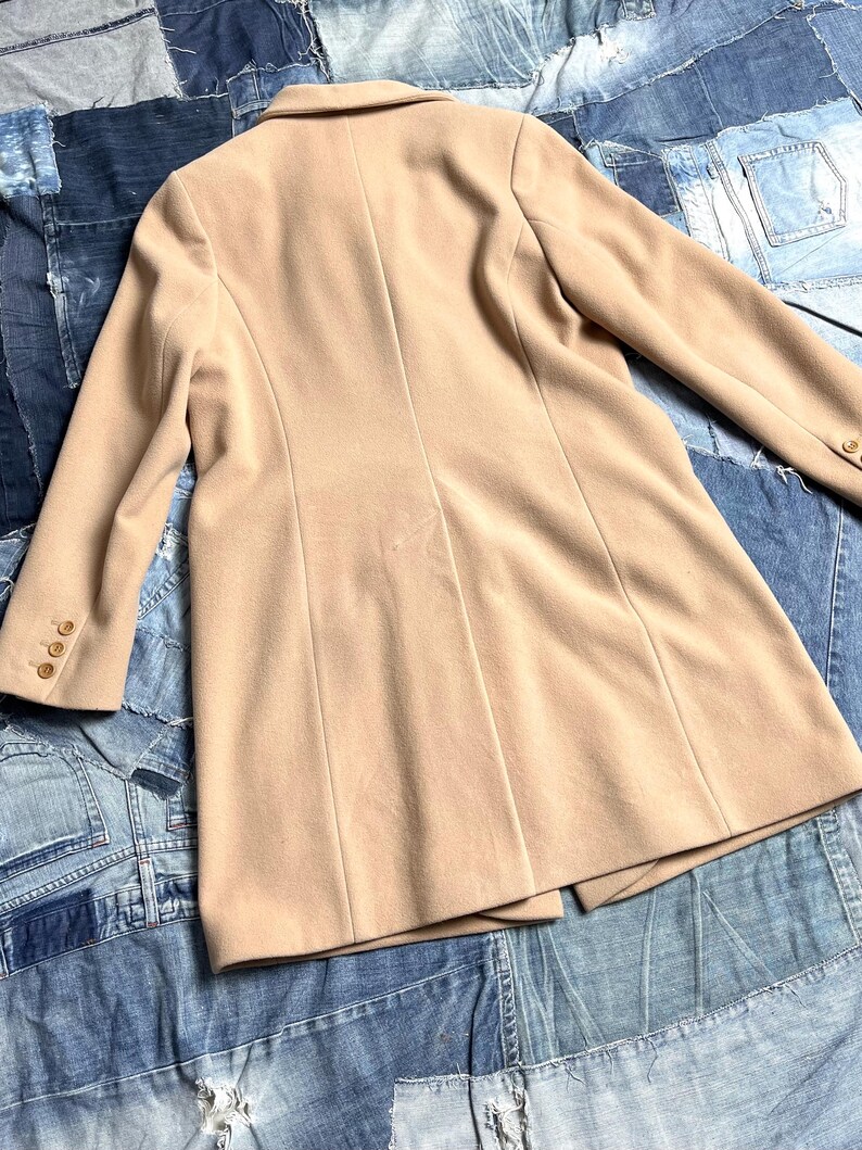 May include: A tan-coloured blazer with a classic design. The blazer features a notched lapel, long sleeves with buttoned cuffs, and a tailored fit. The back of the blazer has vertical seams for a structured look. The garment is made of a smooth fabric.