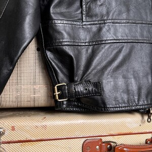 May include: Close-up of a black leather jacket with a buckle detail at the waist. The jacket has a classic design with visible stitching and a textured surface. The jacket is resting on a vintage suitcase.