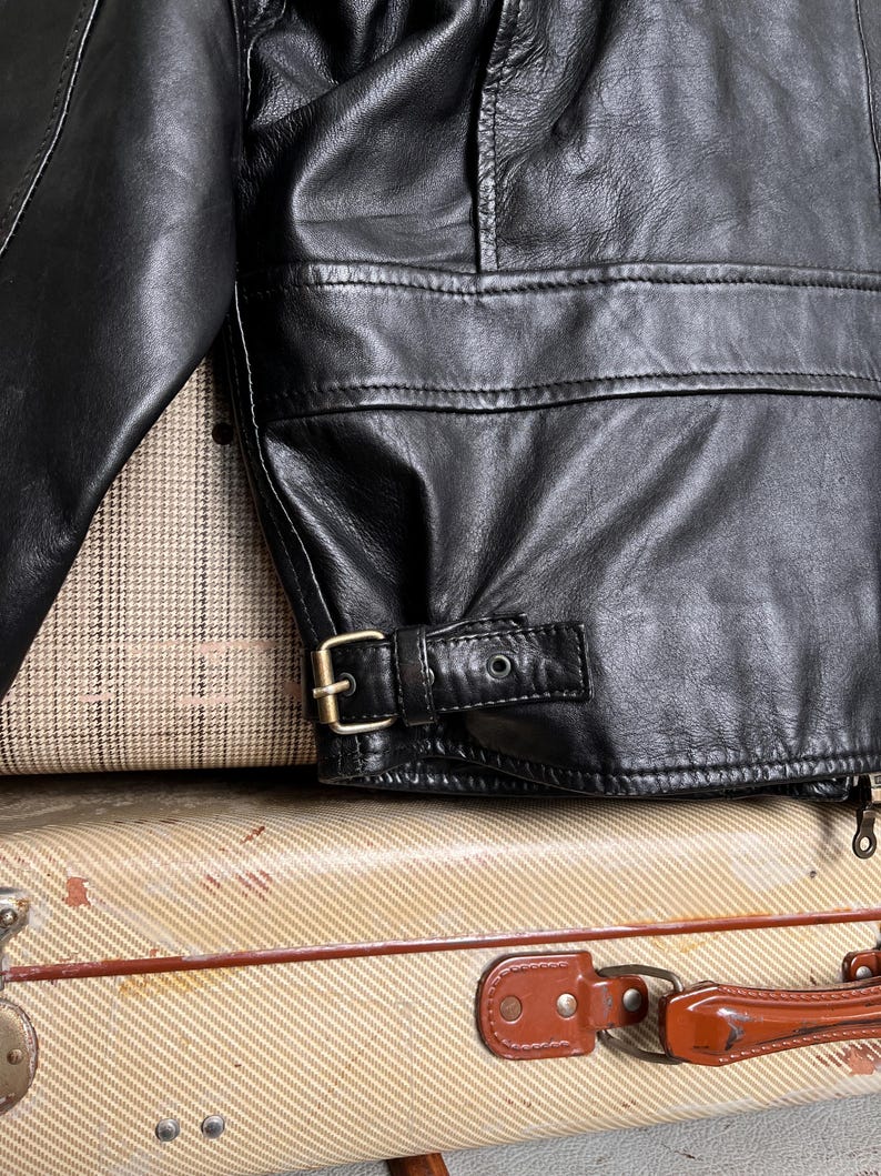 May include: Close-up of a black leather jacket with a buckle detail at the waist. The jacket has a classic design with visible stitching and a textured surface. The jacket is resting on a vintage suitcase.