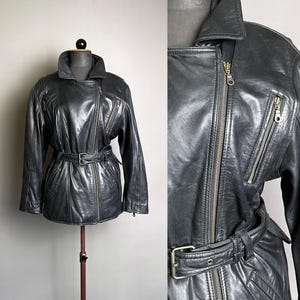 May include: A dark gray leather jacket with a belted waist and asymmetrical zipper closure. The jacket features a notched collar, multiple zippered pockets, and a classic biker style. The jacket is displayed on a mannequin.