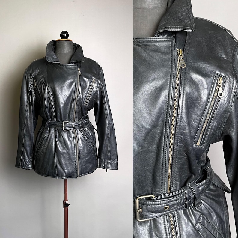 May include: A dark gray leather jacket with a belted waist and asymmetrical zipper closure. The jacket features a notched collar, multiple zippered pockets, and a classic biker style. The jacket is displayed on a mannequin.