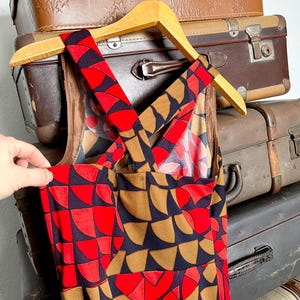 May include: A sleeveless dress featuring a geometric pattern of semicircles in vibrant red, dark navy blue, and muted brown. The garment has wide shoulder straps that cross over the back, creating a distinct crisscross design on the upper bodice.
