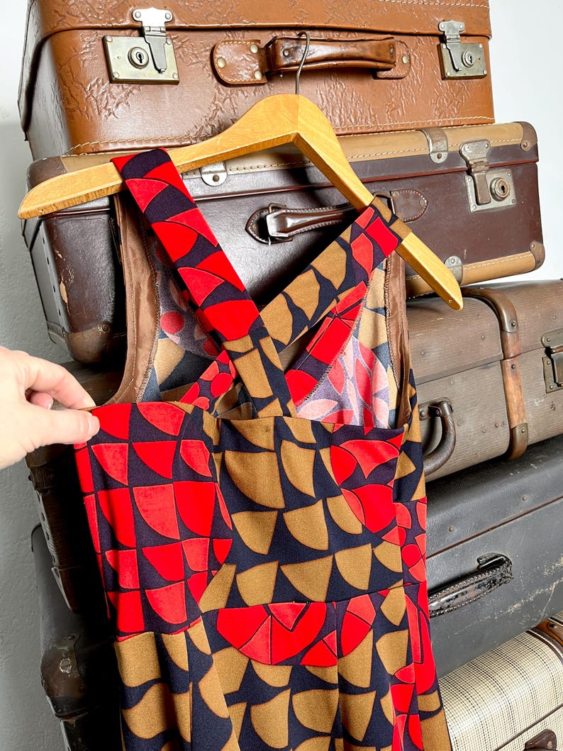 May include: A sleeveless dress featuring a geometric pattern of semicircles in vibrant red, dark navy blue, and muted brown. The garment has wide shoulder straps that cross over the back, creating a distinct crisscross design on the upper bodice.