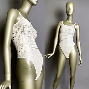 Puede incluir: Un body crema de tejido abierto con tirantes finos. La prenda de una pieza presenta una pernera de corte alto y un patrón texturizado en el corpiño. El body se exhibe en un maniquí sobre un fondo gris.