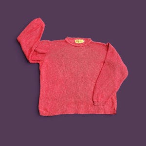May include: A long-sleeved knit sweater in a vibrant coral-pink hue. The garment features a classic crew neckline with a ribbed collar and a small, rectangular yellow brand label stitched inside the neck. The knit texture is consistent throughout.