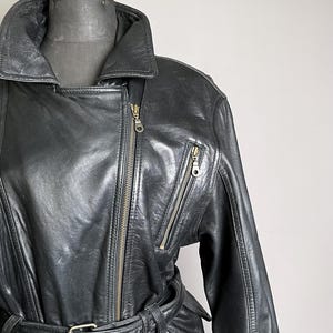 May include: Black leather jacket with an asymmetrical zipper closure, a wide collar, and multiple zippered pockets. The jacket features a belt at the waist and is displayed on a mannequin. The jacket is a classic style.