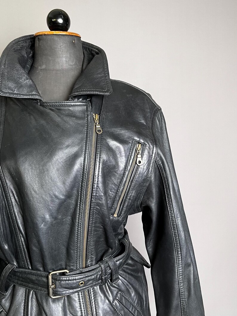 May include: Black leather jacket with an asymmetrical zipper closure, a wide collar, and multiple zippered pockets. The jacket features a belt at the waist and is displayed on a mannequin. The jacket is a classic style.