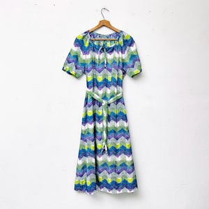 Pode incluir: Um vestido vintage com um padrão geométrico em tons de verde, azul, roxo e branco. O vestido tem uma cintura cintada com um cinto combinando e mangas curtas.