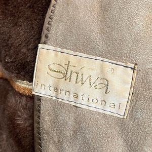 Pu&ograve; includere: Primo piano di una sezione di un capo, forse un cappotto o una giacca, con un materiale strutturato simile alla pelle scamosciata marrone chiaro. Il capo ha una fodera marrone scuro e pelosa. Un'etichetta con la scritta "Striva International" &egrave; cucita.