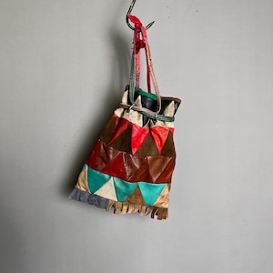Peut inclure: Un sac en cuir patchwork coloré avec fermeture à cordon, suspendu à un crochet métallique. Le sac présente un motif géométrique de triangles rouges, marron, turquoise et crème, avec une frange. Le sac mesure environ 25 cm de long.