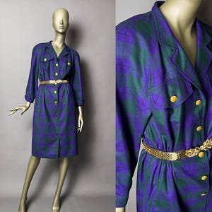 May include: A vintage plaid dress in purple and green, featuring a collared neckline, long sleeves, and gold-tone buttons. The dress is cinched at the waist with a gold belt. The dress is displayed on a mannequin.