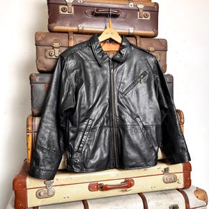 May include: A black leather jacket with a zip-up front and a collared neckline hangs on a wooden hanger. The jacket is displayed on a stack of vintage suitcases in various shades of brown and tan.
