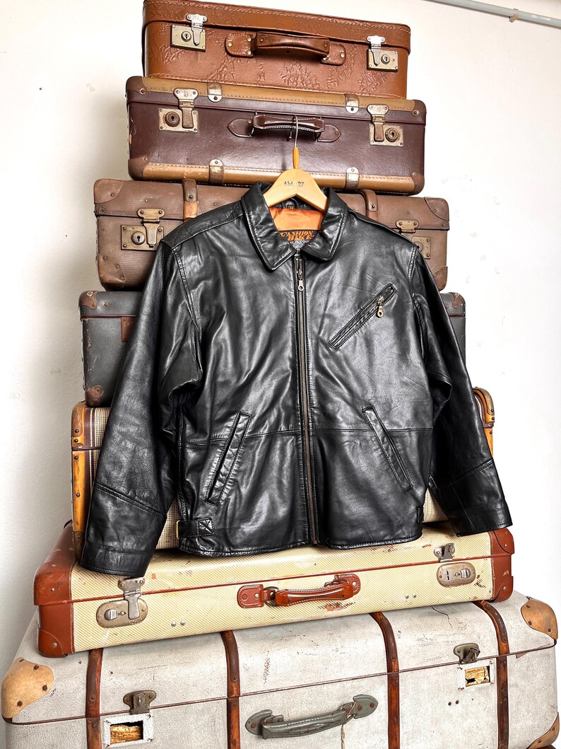 May include: A black leather jacket with a zip-up front and a collared neckline hangs on a wooden hanger. The jacket is displayed on a stack of vintage suitcases in various shades of brown and tan.