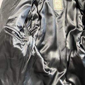 May include: Black leather jacket with a satin lining. The interior features a diamond-shaped tag with a logo. The jacket has a zippered front and a pocket on the left side. The satin lining is a dark gray color.