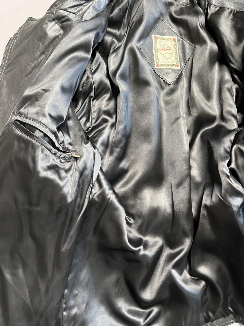 May include: Black leather jacket with a satin lining. The interior features a diamond-shaped tag with a logo. The jacket has a zippered front and a pocket on the left side. The satin lining is a dark gray color.