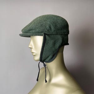 May include: A green tweed flat cap with ear flaps. The cap has a short brim and dark trim around the ear flaps. Black drawstrings are attached to the ear flaps. The hat is displayed on a mannequin head.