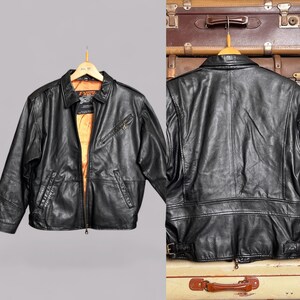 May include: A black leather jacket with a collared neckline, front zipper, and multiple pockets. The jacket has a gold-colored interior lining and is displayed on a wooden hanger. The back of the jacket is also shown, with a belt detail at the waist.