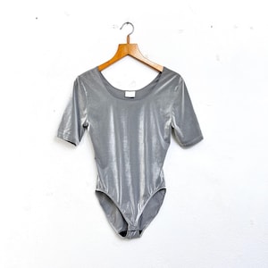 Könnte beinhalten: Ein silberner Metallic-Body mit kurzen Ärmeln und einem runden Ausschnitt. Der Body hängt an einem Holzbügel vor einem weißen Hintergrund.