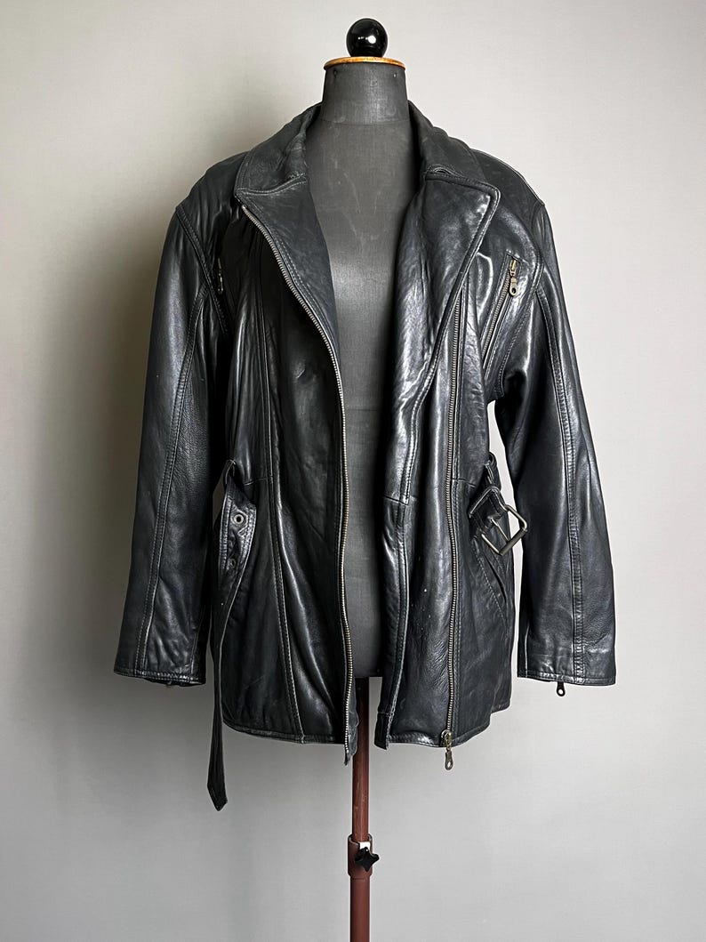 May include: A black leather jacket with a zipper closure, side pockets, and a belt detail. The jacket is displayed on a mannequin against a neutral background. The jacket has a classic biker style.
