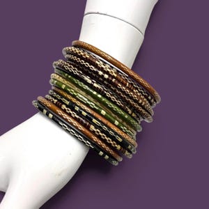 May include: A stack of ten thin bangles featuring a variety of colors including brown, olive green, tan, and black. Each bangle is decorated with a repeating metallic gold rectangular pattern that wraps around the entire circumference of the band.