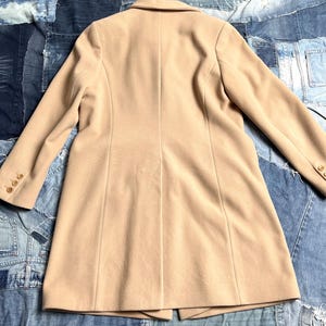 May include: A beige coat with a tailored design. The coat has a notched collar, long sleeves with buttoned cuffs, and a fitted shape. The coat is made of a smooth material and has a single-breasted closure. The coat measures approximately 100 cm in length.