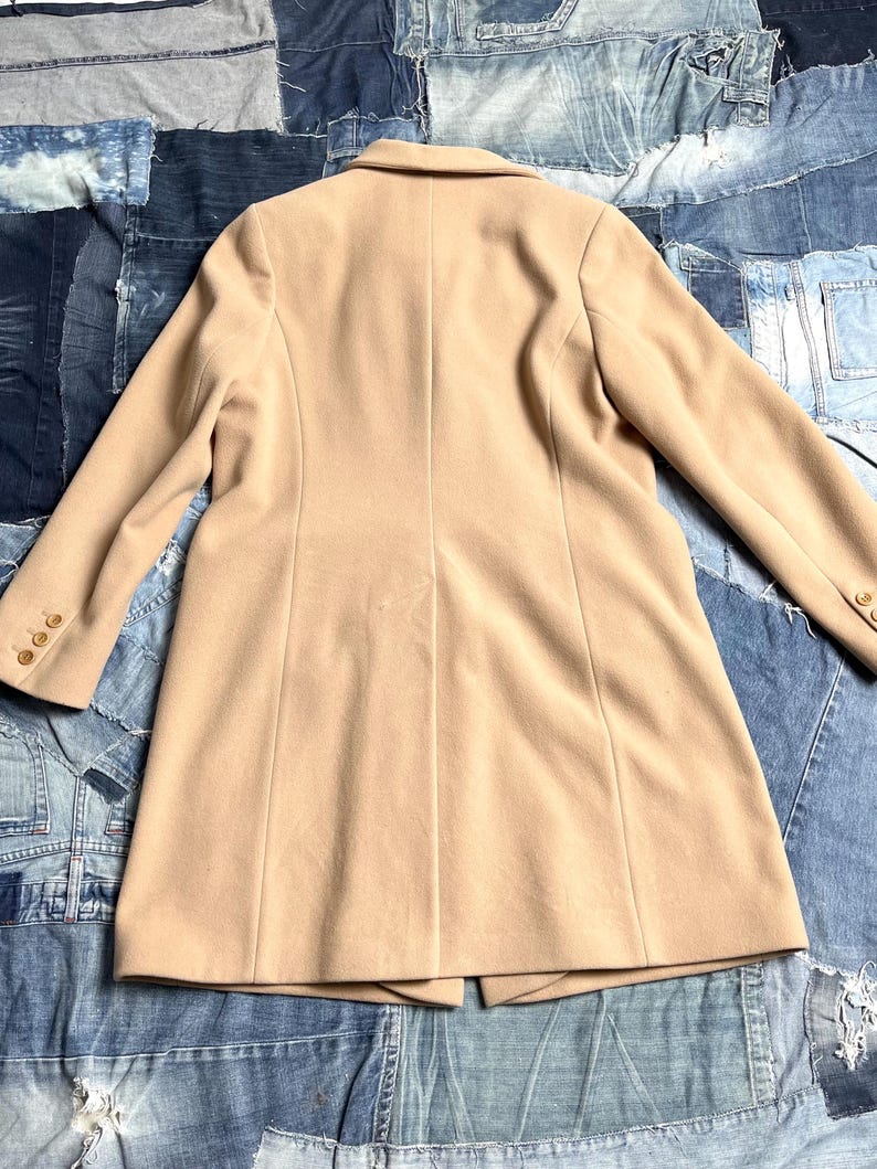 May include: A beige coat with a tailored design. The coat has a notched collar, long sleeves with buttoned cuffs, and a fitted shape. The coat is made of a smooth material and has a single-breasted closure. The coat measures approximately 100 cm in length.