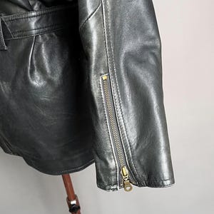 May include: Close-up of a black leather jacket with a zipper detail on the sleeve. The jacket has a classic design with a belt and a textured surface. The zipper is gold-colored.