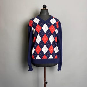 Könnte beinhalten: Ein marineblauer V-Ausschnitt-Pullover mit einem rot, weiß und marinefarbenen Argyle-Muster. Der langärmlige Pullover wird auf einer Schaufensterpuppe vor grauem Hintergrund präsentiert. Das Argyle-Muster ist ein klassisches Design.