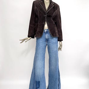 Puede incluir: Una chaqueta de ante marr&oacute;n con costuras blancas y un cierre de un solo bot&oacute;n. La chaqueta se lleva sobre un pantal&oacute;n de mezclilla azul de pierna ancha con dobladillo deshilachado.