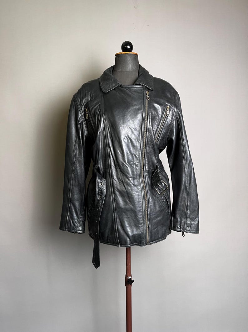 May include: A dark gray leather jacket with an asymmetrical zipper closure, multiple zippered pockets, and a belted waist. The jacket is displayed on a mannequin and has a classic biker style.