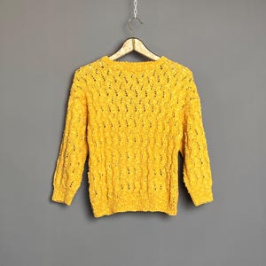 Könnte beinhalten: Ein leuchtend gelber Pullover mit einem strukturierten, offen gestrickten Design. Das langärmlige Kleidungsstück hängt an einem Holzbügel vor einem grauen Hintergrund. Das Strickmuster des Pullovers erzeugt eine visuell interessante Textur.